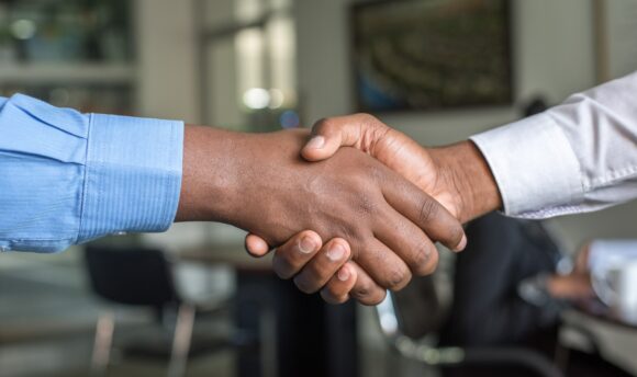 A handshake centered in the image. Sleeve on the left is blue and the skin color of the person is a shade of brown. The sleeve on the right is white and the skin color is light brown. A blurred out office is in the background of the handshake.